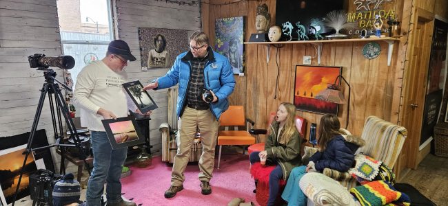 One man holds a large photo, while another man points out aspects of the photo in front of a group of youth.