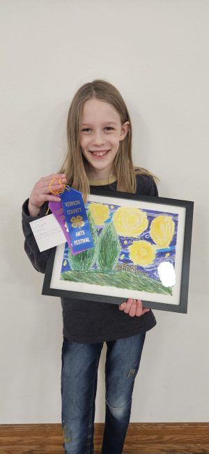 A youth poses with her pencil drawing of a bright sky landscape, and her blue and merit ribbons she received for it.