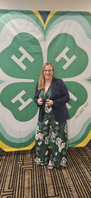4-H Educator, Emery, stands holding a glass award for Distinguished Service.